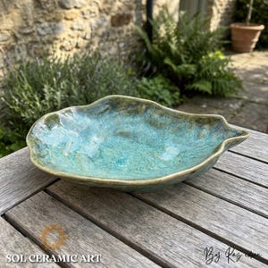 Può includere: Una ciotola artigianale in ceramica a forma di foglia, con smalto turchese e verde. La ciotola ha una superficie strutturata e una forma organica unica. Le parole "SOL CERAMIC ART" sono stampate in basso a sinistra.