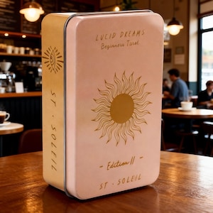 Puede incluir: Una caja metálica rosa con bordes redondeados, con texto dorado y un gráfico de sol. El texto dice "LUCID DREAMS Beginners Tarot". El lateral de la caja es de color beige con el texto "ST. SOLEIL" y un gráfico de ojo.