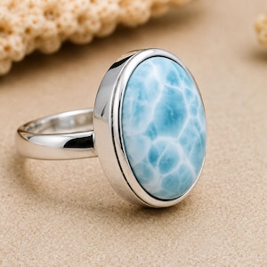 Könnte beinhalten: Ein Ring aus poliertem Silber mit einem großen, ovalen, hellblauen Larimar-Edelstein. Der Stein zeigt ein ausgeprägtes, wolkenartiges Marmormuster in Himmelblau und Weiß und ist sicher in einer glatten, massiven Silberschiene gefasst.