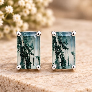 May include: A pair of rectangular stud earrings featuring moss agate gemstones with deep green, tree-like dendritic inclusions set in polished silver prongs. The stones are translucent with a smooth, faceted surface, showcasing natural patterns.