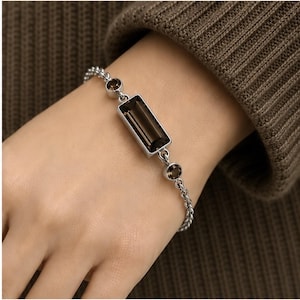 May include: A silver-toned chain bracelet featuring a central rectangular smoky quartz gemstone in a bezel setting, flanked by two smaller round smoky quartz stones. The bracelet is constructed with a delicate metal link chain and secure metal findings.