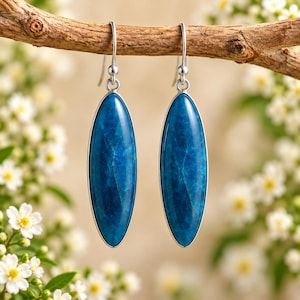 Puede incluir: Un par de pendientes largos de forma ovalada con piedras lisas de color azul intenso y vetas internas sutiles. Las piedras están engastadas en un fino marco de metal plateado y cuelgan de sencillos ganchos de metal plateado.