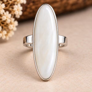 May include: A silver-toned ring featuring a large, elongated oval-shaped white stone set vertically. The stone has a smooth, pearlescent surface with subtle cream-coloured marbling, held within a polished metal bezel and a simple, solid band.