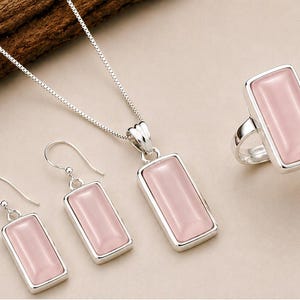 Op de afbeelding: Een sieradenset bestaande uit een zilveren ketting met een rechthoekige hanger, een paar bijpassende rechthoekige oorbellen en een rechthoekige ring, allemaal voorzien van gladde, lichtroze cabochonstenen in zilveren monturen.
