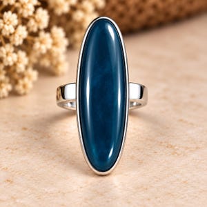 Può includere: Un anello in argento con una grande pietra ovale di colore blu scuro, liscia e montata verticalmente. La fascia in metallo lucido sostiene il cabochon allungato, che presenta un colore blu profondo e uniforme con sottili variazioni interne e una finitura lucida.