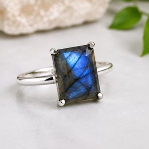 Puede incluir: Un anillo de tono plateado con una gran piedra preciosa de labradorita rectangular y facetada. La piedra muestra un destello azul iridiscente prominente en su superficie y está asegurada por cuatro garras metálicas en una banda fina, lisa y pulida.