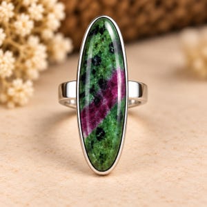 Può includere: Un anello in argento con un grande cabochon ovale allungato in zoisite con rubino. La pietra presenta una base screziata verde e nera con una prominente striscia diagonale di colore magenta intenso che attraversa il centro della montatura.