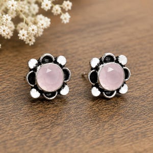 Puede incluir: Un par de pendientes de botón con una piedra central redonda facetada de color rosa pálido, engastada en un marco de plata oxidada oscura con forma de flor de ocho pétalos redondeados. El metal tiene un acabado de alto contraste con zonas oscuras.