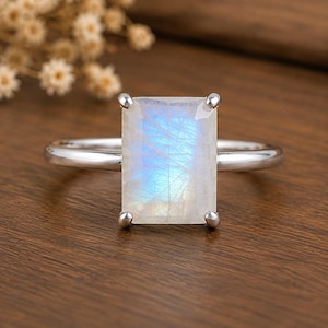May include: A silver-toned ring featuring a large, rectangular, faceted moonstone centre gem. The stone displays a translucent, milky white appearance with a distinct blue adularescence glow. The gem is secured by four metal prongs on a band.