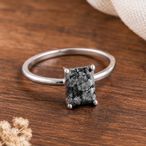 Puede incluir: Un anillo de plata con una piedra rectangular facetada de patrón moteado en negro y gris. La piedra está sujeta por cuatro pequeñas garras metálicas sobre una banda de plata lisa, fina y pulida que forma una figura circular completa.