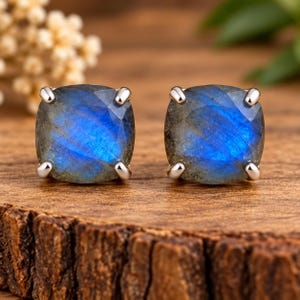 Puede incluir: Un par de pendientes de botón con piedras de labradorita de corte cuadrado y superficie facetada. Cada piedra muestra una iridiscencia azul vibrante y está asegurada por cuatro garras de metal plateado, creando un aspecto pulido y brillante.
