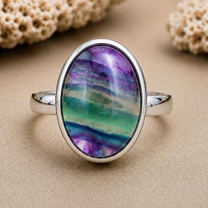 Puede incluir: Un anillo de plata con una gran piedra preciosa pulida de forma ovalada, que presenta bandas horizontales de colores violeta vibrante, azul profundo y verde brillante. La piedra está engastada en un bisel de plata lisa y pulida.