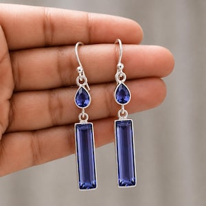 May include: A pair of silver drop earrings featuring a small teardrop-shaped blue gemstone suspended above a larger rectangular blue gemstone. The earrings are attached to silver ear wires and have a polished metallic finish with deep blue stones.