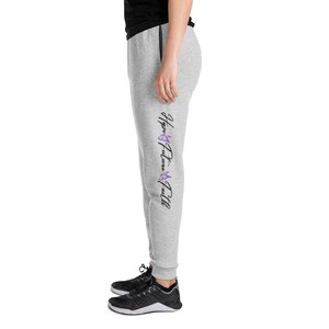 Peut inclure: Pantalon de survêtement gris clair avec les mots "Hope Future Faith" imprimés verticalement en script noir avec des accents violets. Le pantalon de survêtement a un cordon de serrage noir et une bordure noire le long des poches. La personne porte des chaussures de sport noires.