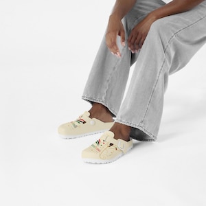 Puede incluir: Zuecos sin cordones de color crema con un estampado gráfico de una cabeza de elefante estilizada con la trompa curvada hacia arriba, acentuado por rayas verdes, blancas y rojas, pequeños corazones rojos y el texto "Ganesh" en letra cursiva.