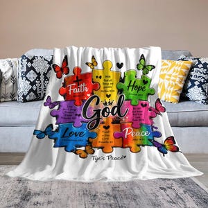 May include: A white throw blanket featuring a colourful puzzle piece design with the word God in the centre. The puzzle pieces include the words Faith, Hope, Love, and Peace, along with various Bible verses and several butterflies in different colours.