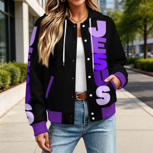 May include: A black varsity-style hooded jacket featuring white drawstrings and a snap-button front closure. The jacket displays the word JESUS in large, vertical, light purple block lettering on both sides of the front, with purple cuffs.
