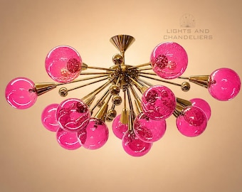 May include: A modern chandelier featuring multiple pink glass globes and gold-colored metal arms. The fixture has a central hub with radiating arms, each ending in a pink globe. The text "LIGHTS AND CHANDELIERS" is visible.