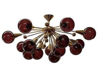 May include: A mid-century modern chandelier featuring a brass frame with multiple arms extending outwards. Each arm holds a round, dark red glass globe. The fixture has a central cone-shaped element and smaller brass spheres.