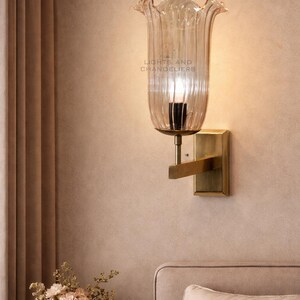 May include: A wall sconce with a clear, tulip-shaped glass shade and a brass-colored metal base. The sconce is mounted on a beige wall. The text "LIGHTS AND CHANDELIERS" is visible on the glass. A vase of flowers sits on a nearby surface.