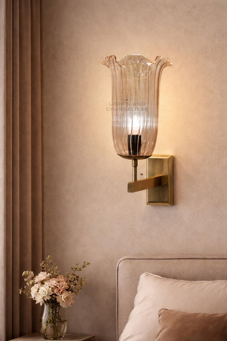 May include: A wall sconce with a clear, tulip-shaped glass shade and a brass-colored metal base. The sconce is mounted on a beige wall. The text "LIGHTS AND CHANDELIERS" is visible on the glass. A vase of flowers sits on a nearby surface.