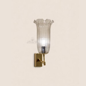 May include: A wall sconce with a clear, fluted glass shade shaped like a flower. The sconce has a gold-colored metal base and arm. The text "LIGHTS AND CHANDELIERS" is visible on the wall.