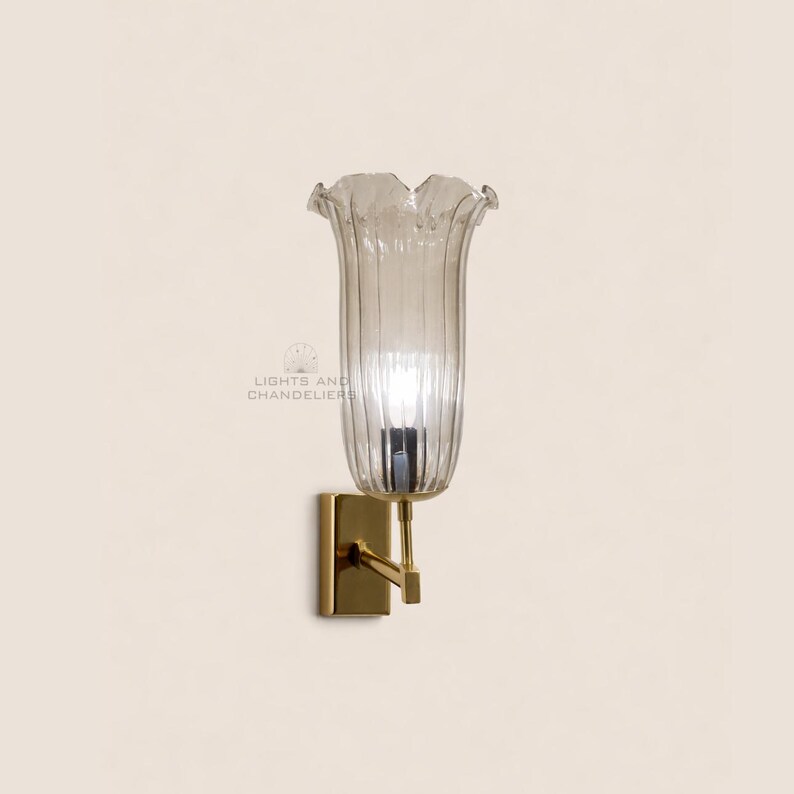 May include: A wall sconce with a clear, fluted glass shade shaped like a flower. The sconce has a gold-colored metal base and arm. The text "LIGHTS AND CHANDELIERS" is visible on the wall.