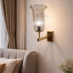 May include: A wall-mounted light fixture with a clear, fluted glass shade shaped like a flower. The fixture has a gold-colored arm and a rectangular base. The text "LIGHTS AND CHANDELIERS" is visible on the shade.