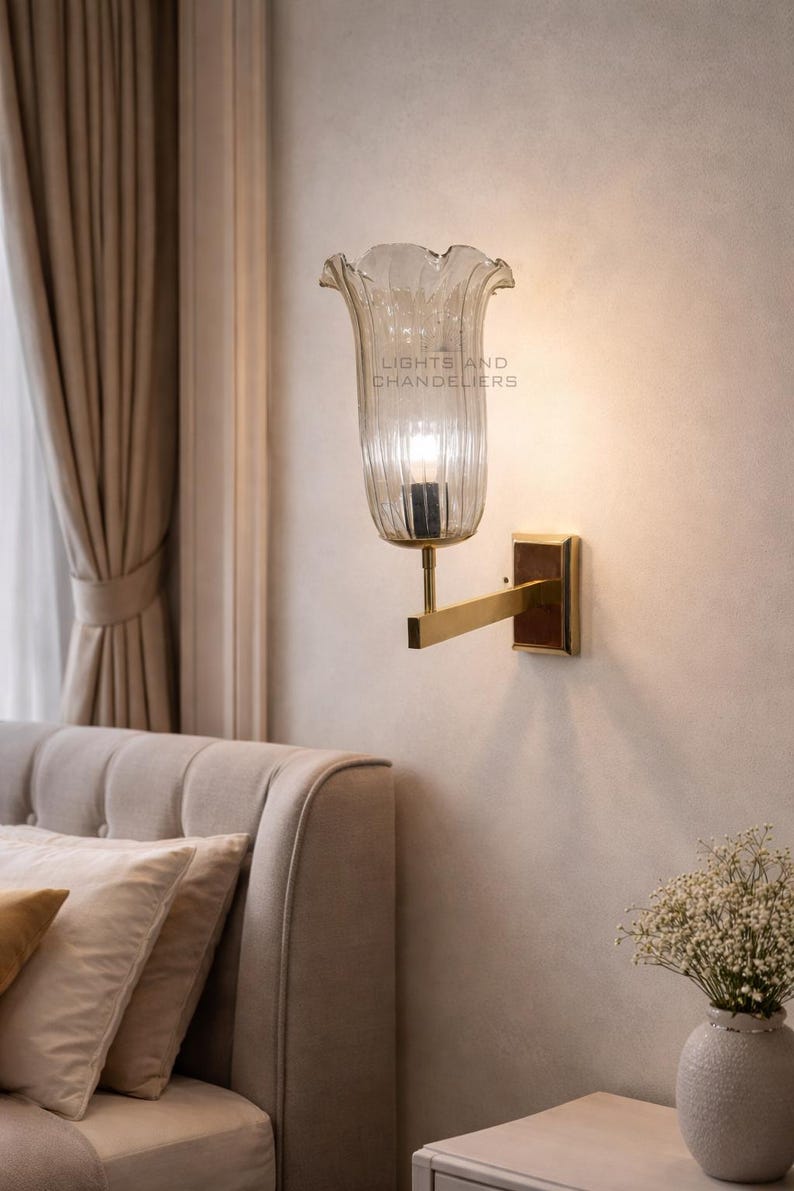 May include: A wall-mounted light fixture with a clear, fluted glass shade shaped like a flower. The fixture has a gold-colored arm and a rectangular base. The text "LIGHTS AND CHANDELIERS" is visible on the shade.