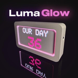 Op de afbeelding: Een wit digitaal display met de tekst "OUR DAY" en het nummer "36" in roze. Het display heeft een roze rand en de merknaam "Luma Glow" staat erboven. De weergave wordt hieronder weerspiegeld.