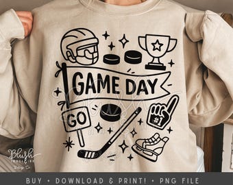 May include: Beige sweatshirt with a black graphic design featuring hockey-related elements. The design includes a hockey helmet, pucks, a trophy, a hockey stick, a foam finger, a skate, and the words "GAME DAY" and "GO".