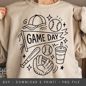 May include: Beige sweatshirt featuring a black baseball-themed design. The graphic includes a baseball cap, baseballs, a bat, a glove, a drink cup, and the words "GAME DAY" on a banner. Stars and lines complete the design.