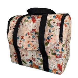 Può includere: Una borsa da viaggio con stampa floreale con sfondo pesca e fiori colorati in rosso, blu e arancione. La borsa ha manici neri e una cerniera nera. La borsa è rettangolare e ha una maniglia superiore.