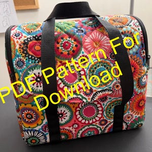 Op de afbeelding: Een kleurrijke gewatteerde tas met een levendig bloemenpatroon in tinten rood, oranje, blauw en groen. De tas heeft zwarte handvatten en de tekst "PDF Pattern For Download" in geel.