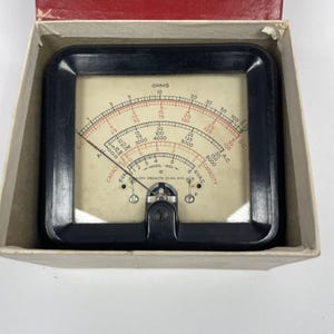 May include: Vintage black and cream-colored electrical meter with a needle and multiple scales for measuring ohms, voltage, and capacity. The meter is housed in a cardboard box with a red interior.