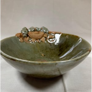 May include: A handmade ceramic bowl with a wide, shallow design. The bowl has a green and brown glaze with a decorative relief of small, round shapes and leaves on the rim. The bowl is suitable for serving food or as a decorative piece.