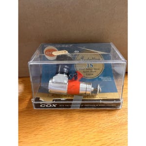 May include: A Cox Medallion .15 front rotary valve sport-stunt engine encased in a clear plastic display box. The engine features a silver metal body with a bright orange section and a black cylinder head. The box includes branding and text.