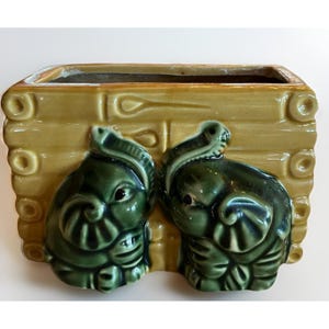 May include: A rectangular ceramic planter with a light yellow-brown exterior and a dark green interior. Two green elephant figures are on the front. The planter has a textured, log-cabin-like design.