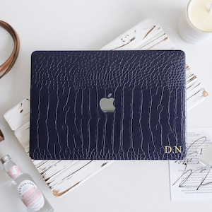 May include: A dark navy blue laptop case with a textured crocodile skin pattern and a silver Apple logo in the center. The bottom right corner features the gold-colored initials D.N. printed on the surface of the protective cover.