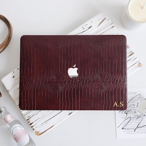 May include: A dark burgundy laptop case with a textured crocodile skin pattern and a cutout for the Apple logo. The bottom right corner features the gold-colored initials A.S. The case is rectangular with rounded corners and a smooth finish.