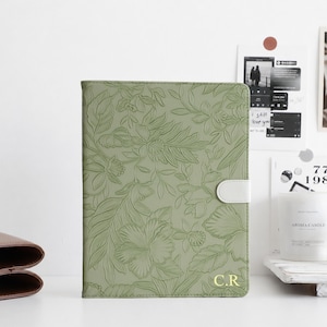 May include: A sage green notebook cover featuring an embossed floral pattern and a white magnetic closure tab. The bottom right corner displays the gold-coloured initials C.R. The cover has a rectangular shape with stitched edges.