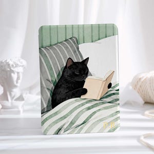 May include: A rectangular print featuring a black cat reading a book in bed. The cat is nestled among pillows and a striped blanket in shades of green and gray. The book has beige pages.