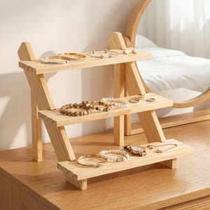 Puede incluir: Un expositor de joyas de madera de tres niveles con varias pulseras, anillos y un reloj. El expositor es de madera de color claro y está colocado sobre una cómoda de madera. Un espejo redondo con marco de madera está en el fondo.