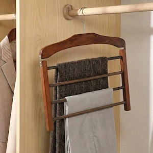 Puede incluir: Un colgador de pantalones de madera de varios niveles con tres barras horizontales, que sostiene un par de pantalones grises y un par de pantalones de tweed marrones. El colgador está suspendido de un gancho de metal sujeto a una barra de madera dentro de un armario.