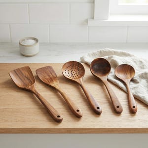 May include: A collection of five wooden kitchen utensils, including a spatula, slotted spoon, and various spoons. The utensils are made of a warm-toned wood and are arranged on a light-colored wooden surface.
