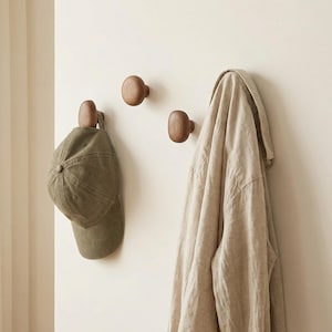 May include: Three wooden wall hooks in a light-coloured room. One hook holds a green baseball cap, and another holds a beige jacket. The hooks are a warm brown colour and are mounted on a neutral-toned wall.