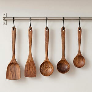 May include: A set of five wooden kitchen utensils hanging from a metal bar. The utensils include a spatula, a flat turner, a slotted spoon, a rounded spoon, and a small ladle. The wood grain is visible, and the color is a warm brown.