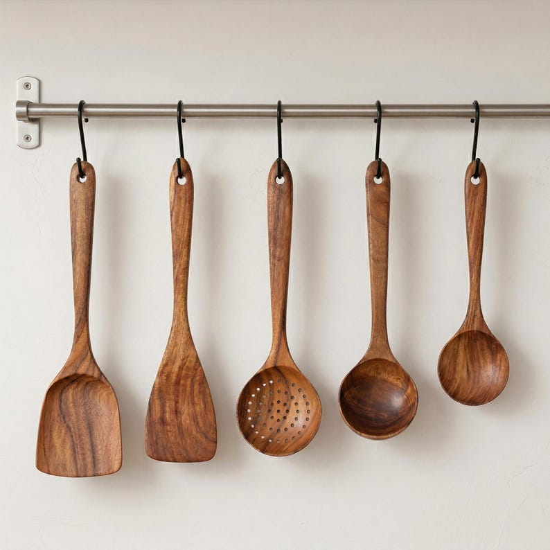 May include: A set of five wooden kitchen utensils hanging from a metal bar. The utensils include a spatula, a flat turner, a slotted spoon, a rounded spoon, and a small ladle. The wood grain is visible, and the color is a warm brown.