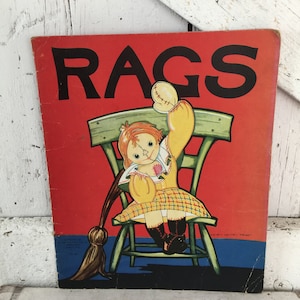 May include: Vintage book cover with the word "RAGS" in large black letters on a red background. The illustration features a doll sitting on a green chair, wearing a yellow dress and holding a broom.