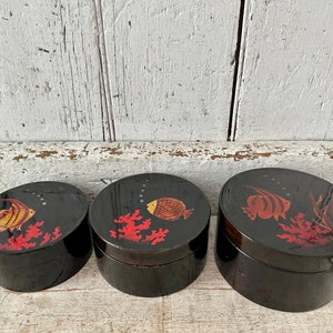 May include: Set of three black, oval-shaped boxes with lids. Each box features a different fish design in shades of gold and red, along with red coral accents. The boxes are arranged in a nested configuration.